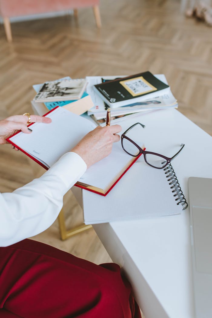 A Person Writing on a Notebook Near Eyeglasses